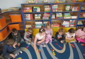 Dzieci siedzą na dywanie i układają obrazki z figur geometrycznych.