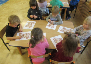 Dzieci siedzą przy stolikach i wyklejają obrazki misiów plasteliną.
