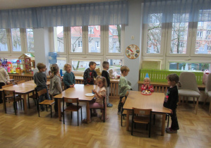 Świętujemy urodziny dziewczynki, jubilatka siedzi przy stoliku w urodzinowej czapeczce, pozostałe dzieci stoją przy stolikach i śpiewają "Sto lat".