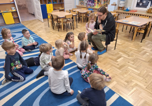 Dziewczynka z tatą siedzą na krzesełkach, tata czyta książkę, pozostałę dzieci siedzą na dywanie i słuchają.