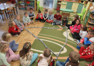 Dzieci siedzą w kole, przed nimi trójkąt ułożony z łańcuchów świątecznych.