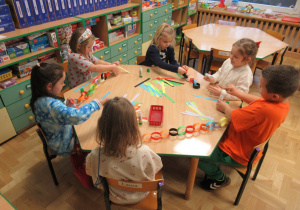 Dzieci siedzą przy stole i kleją łańcuchy z kolorowego papieru.