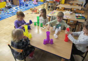 Dzieci siedzą przy stoliku i układają kolorowe kubeczki.