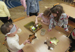 Dziewczynka z zasłoniętymi oczami wącha cynamon, dziewczynki dotykają wacików i szyszek umieszczonych w pudełku.