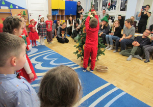 Chłopiec wiesza gwiazdkę na choince, rodzice oglądają występ świąteczny dzieci.