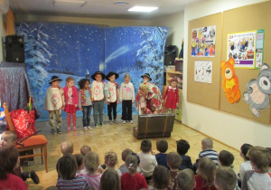 Kilkoro dzieci stoi w szerego, pani w stroju góralskim wyjmuje coś z walizki, pozostałe dzieci siedzą na podłodze i obserwują.