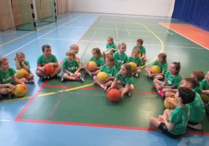 Dzieci w strojach gimnastycznych siedzą na podłodze w sali gimnastycznej, niektóre dzieci trzymają piłkę.