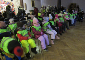Dzieci siedzą na krzesłach i czekają na przedstawienie wystawiane przez seniorów.