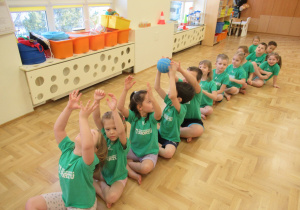 Dzieci siedzą w rzędzie i podają sobie piłkę nad głowami.