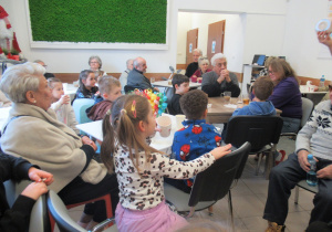 Dzieci siedzą przy stolikach z Seniorami i piją herbatkę.