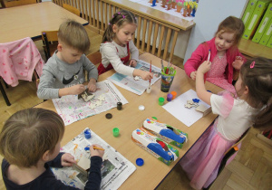 Dzieci przy stoliku malują farbami kwiatki wykonane z drewna.