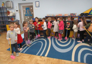 Dzieci stoją w półkolu, trzymają w rękach "miotełki".