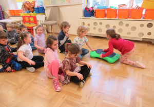 Dzieci siedzą w dwóch rzędach, dziewczynka podaje chłopcu jeździk gimnastyczny.