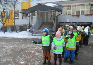 Dzieci stoją w kamizelkach odblaskowych przed szkołą.