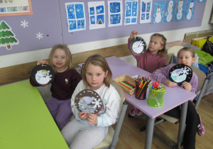 Dziewczynki siedzą w ławkach w szkole i prezentują wykonane na zajęciach prace plastyczne.