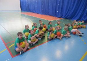 Dzieci siedzą na podłodze na sali gimnastycznej, każde dziecko trzyma piłkę.