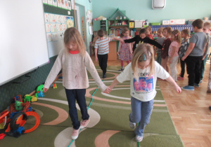 Dzieci przechodzą po linach ułożonych na dywanie trzymając się za rękę.