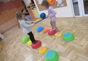Dziewczynki przechodzą po kamieniach sensorycznych i przenoszą piłkę na laskach gimnastycznych.