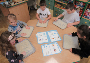 Dzieci siedzą przy stoliku i piszą litery na tackach z ryżem.