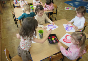 Dzieci siedzą przy stolikach i malują farbami serca.