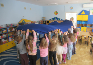 Dzieci stoją w kole i trzymają nad sobą niebieski materiał.