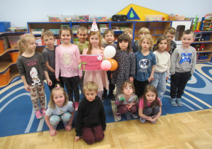 Świętujemy urodziny dziewczynki, jubilatka ma na głowie papierową czapeczkę, wokół stoją inne dzieci.