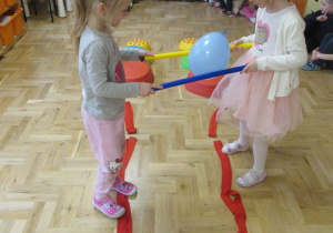 Dwie dziewczynki przenoszą balon, który trzymają między dwoma laskami gimnastycznymi.