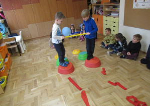 Dwaj chłopcy przenoszą balon, który trzymają między dwoma laskami gimnastycznymi, chłopcy przechodzą po kamieniach sensorycznych.
