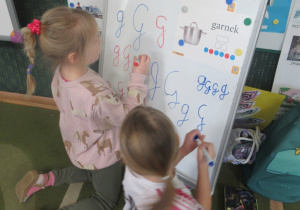 Dziewczynki piszą literę g na tablicy.
