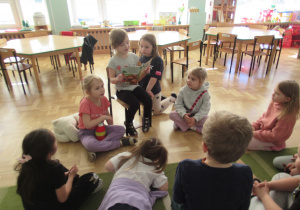 Dziewczynka siedzi na krześle i czyta książkę, pozostałe dzieci siedzą na podłodze i słuchają.