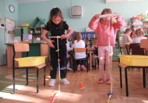 Dwie dziewczynki stoją, trzmają w rękach rolki papierowe i nawijają na nie wstążki.