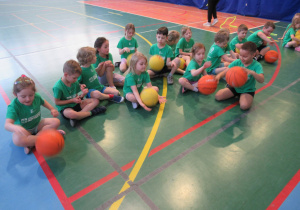 Dzieci siedzą na podłodze trzymając piłki do koszykówki w sali gimnastycznej.