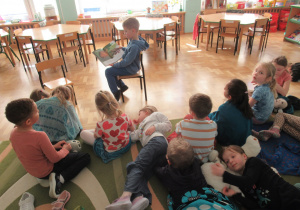 Chłopiec siedzi na krześle i czyta książkę, pozostałe dzieci siedzą na dywanie i słuchają.