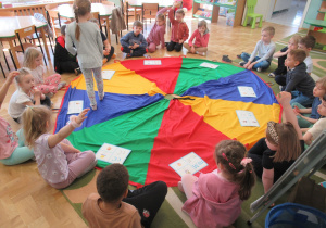 Ddzieci siedzą wokół chusty animacyjnej, dziewczynka stoi na chuście.