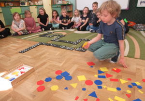 Chłopiec trzyma w ręku figurę geometryczną, na podłodze leżą figury geometryczne.