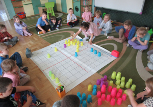 Dzieci siedzą wokół maty do kodowania, dziewczynka kładzie kolorowy kubek na macie.