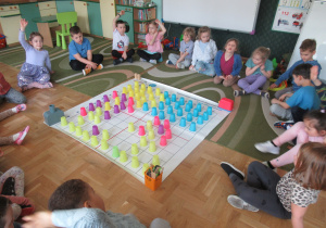 Dzieci siedzą wokół maty do kodowania, na macie ułożony jest obrazek z kolorowych kubeczków.