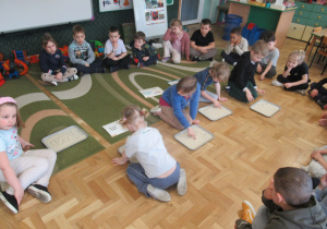 Dzieci siedzą w kole, kilkoro dzieci pisze litery w na tackach wysypanych ryżem.