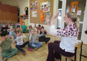 Dzieci siedzą na podłodze z uniesionymi rękami, przed nimi pani od rytmiki.