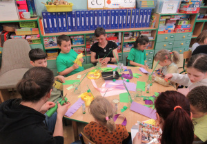 Dzieci z rodzicami siedzą przy stole i wykonują kwiaty z papieru podczas warsztatów wiosennych.