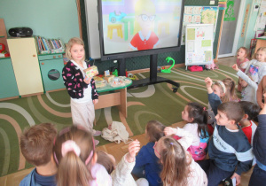 Dziewczynka trzyma w ręku produkt ze znaczkiem ekologicznym, pozostałe dzieci siedzą na podłodze.